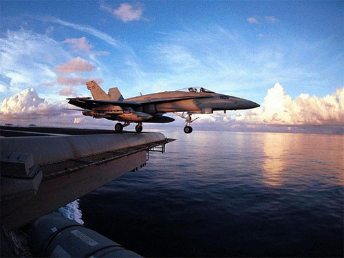 An F/A-18 Hornet alongside an aircraft carrier at sunset