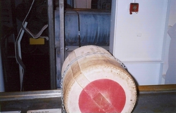 One powder bag on a floor-mounted conveyor inside the USS North Carolina powder magazine, with chain barrier and red ignition pad