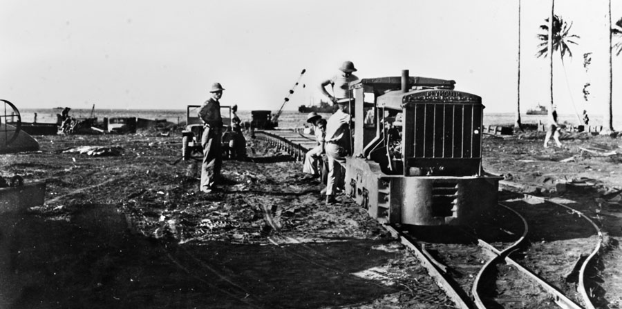 Seabees laying narrow-gauge track across a beachhead with supply ships offshore