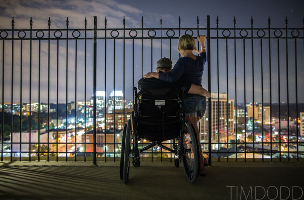 Taylor Morris at a railing above a city skyline at night, his wife sitting beside him with an arm around his shoulders