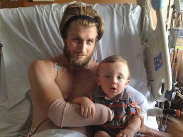 Taylor Morris sitting up in his hospital bed holding a small child, his residual limbs still bandaged